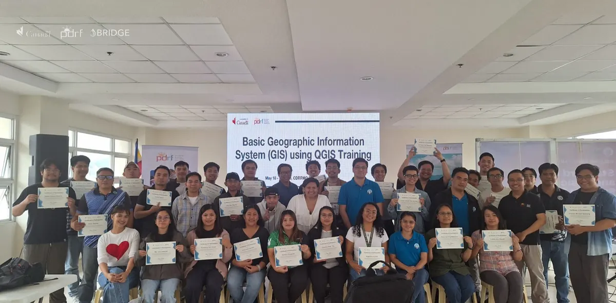 Participants holding their certificates of participation in front of a screen reading \u201cBasic Geographic Information System (GIS) using QGIS Training\u201d.